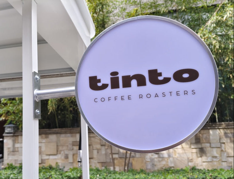 Round sign for tinto Coffee Roasters outside a building with greenery in the background.